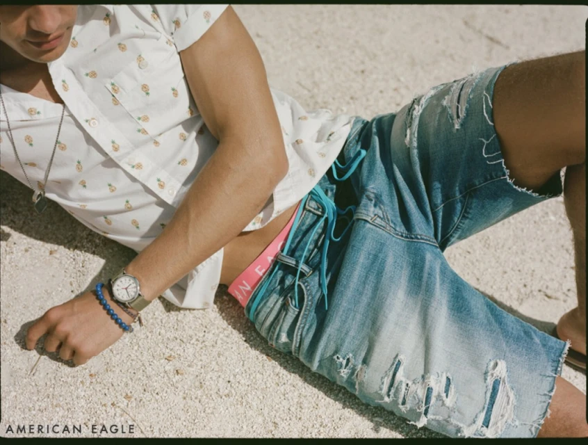 A young adult man poses on a sandy surface wearing a patterned short-sleeve shirt and distressed denim shorts, showcasing casual style.