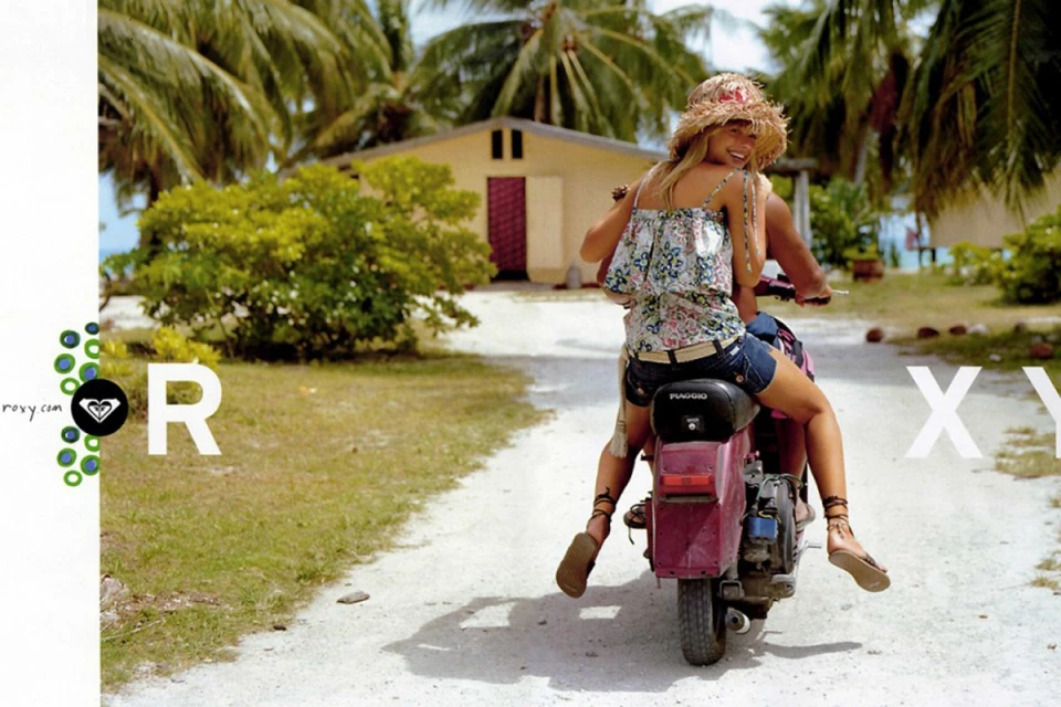 Two women ride a pink moped down a sandy path lined with palm trees and green foliage in a tropical outdoor setting on a sunny day.