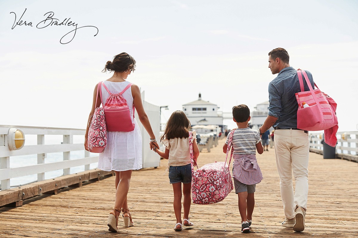 Family of four walks on a pier with pink Vera Bradley bags. The mother carries a backpack, the father a tote, and the children carry duffel bags.