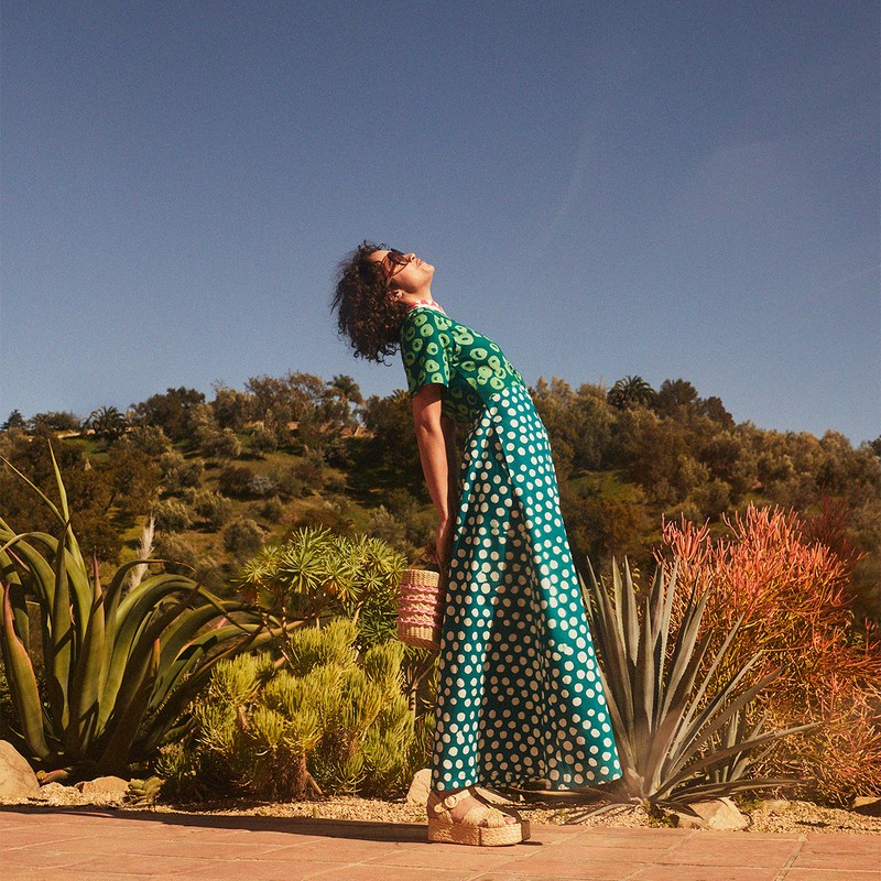 Woman in a patterned green dress and platform shoes smiles with her head tilted back in an outdoor desert garden setting.