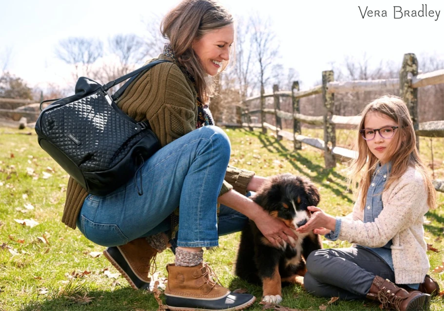 A woman and a child petting a Bernese Mountain Dog puppy on a grassy lawn. A black quilted Vera Bradley bag sits on the grass nearby.