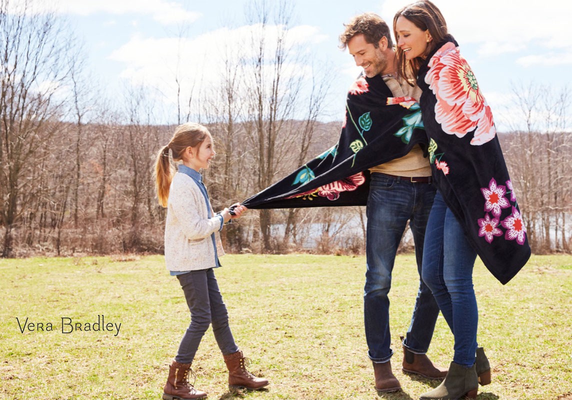 Outdoor family portrait: A man and woman are wrapped in a floral blanket while a young girl pulls at the blanket's edge, all smiling.