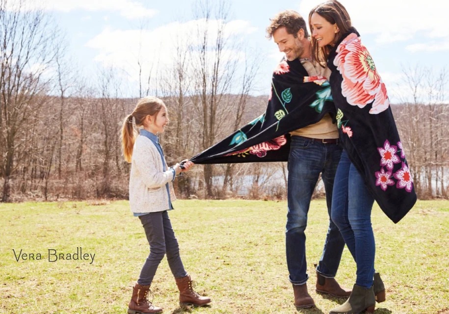 Outdoor family portrait: A man and woman are wrapped in a floral blanket while a young girl pulls at the blanket's edge, all smiling.