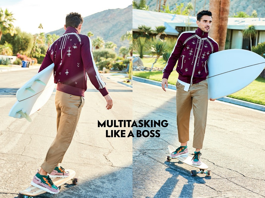 Man in burgundy jacket and khaki pants on skateboard holding surfboard. Outdoor scene with mountains and Palm Springs-style architecture.