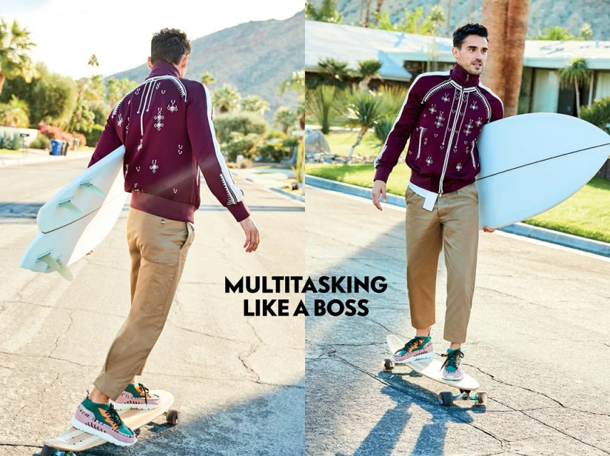 Man in burgundy jacket and khaki pants on skateboard holding surfboard. Outdoor scene with mountains and Palm Springs-style architecture.