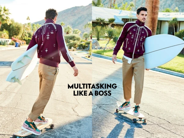 Man in burgundy jacket and khaki pants on skateboard holding surfboard. Outdoor scene with mountains and Palm Springs-style architecture.