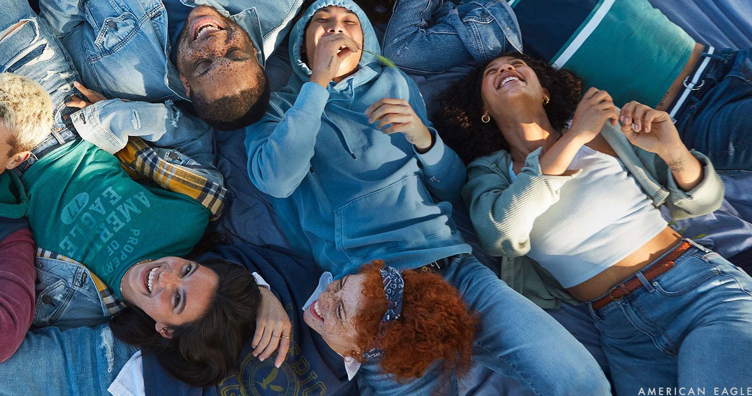Diverse group of smiling young adults lying together on blue material, wearing denim jackets, jeans, and casual tops in an outdoor setting.