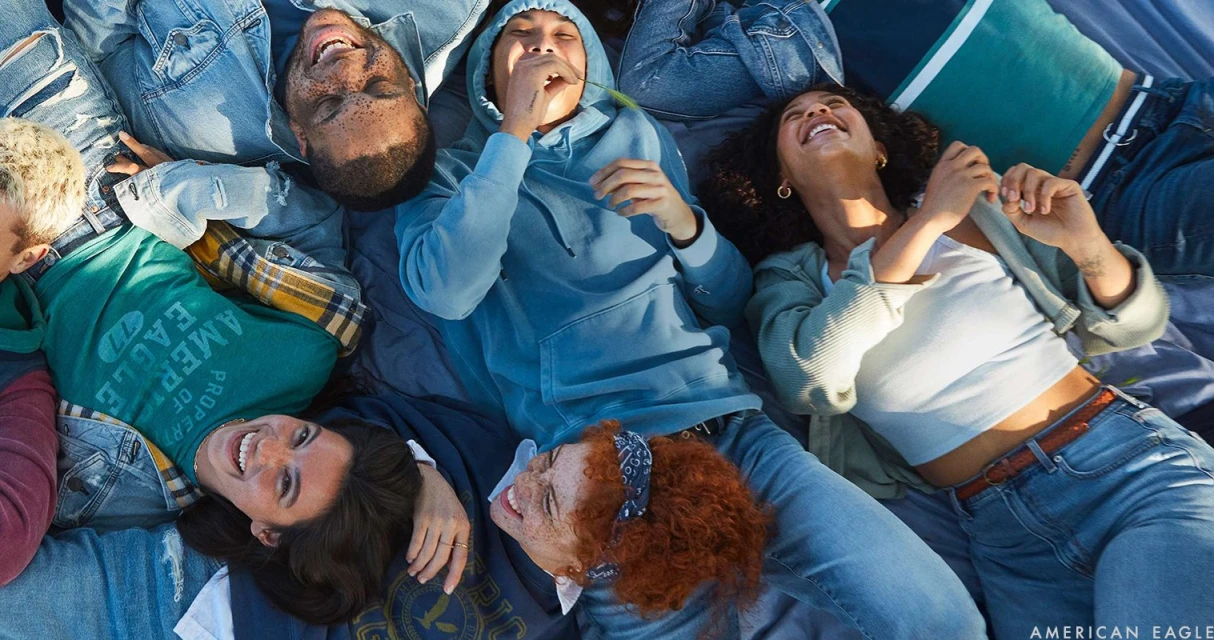 Diverse group of smiling young adults lying together on blue material, wearing denim jackets, jeans, and casual tops in an outdoor setting.