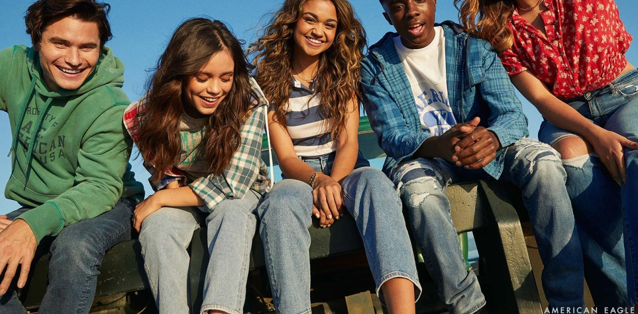Group of five young adults smiling and sitting together outside, wearing casual clothing such as jeans, hoodies, and floral tops in daylight.
