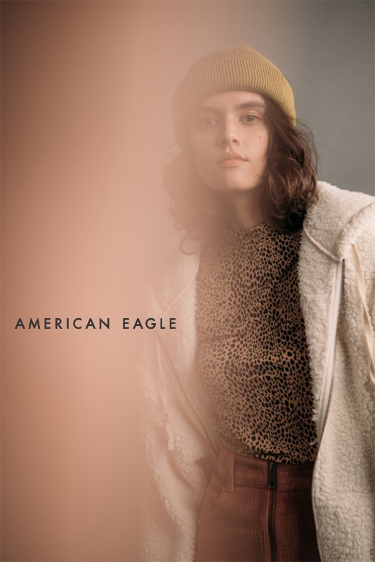 Fashion portrait of a young adult woman wearing a beanie, animal-print shirt, corduroy pants, and jacket with the American Eagle logo.