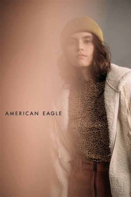 Fashion portrait of a young adult woman wearing a beanie, animal-print shirt, corduroy pants, and jacket with the American Eagle logo.