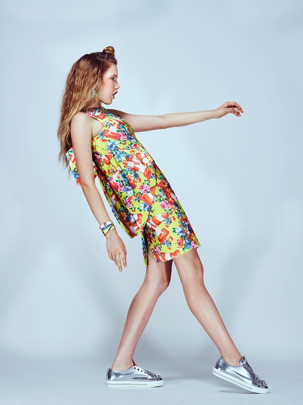 Young woman in a short floral dress poses sideways in shiny shoes against a light background in a fashion studio shot, arms outstretched.