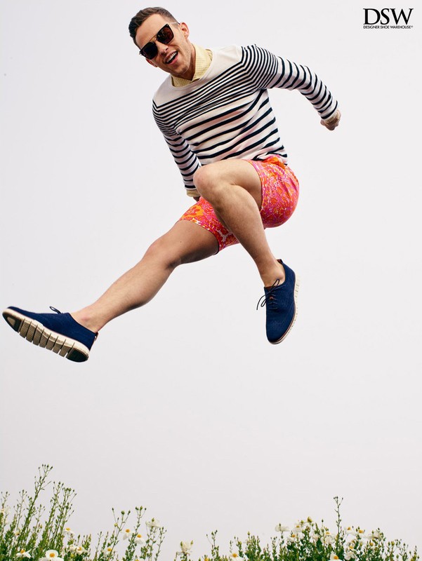 Adult male in a striped sweater and colorful shorts leaps in the air wearing blue shoes, photographed against a bright sky and flowers.