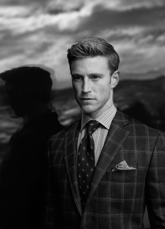 Black and white portrait of a blond-haired, adult male wearing a patterned suit, tie, and pocket square, against a cloudy background.
