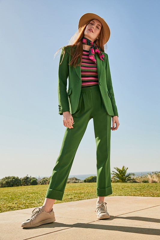 Full-body shot of a woman in a green suit and straw hat standing outside on a sunny day. Fashion editorial with outdoor backdrop.