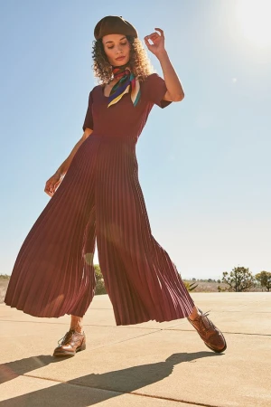 Model in pleated burgundy jumpsuit wearing a beret and colorful scarf outdoors under a clear sky, styled by Eddie Schachnow.