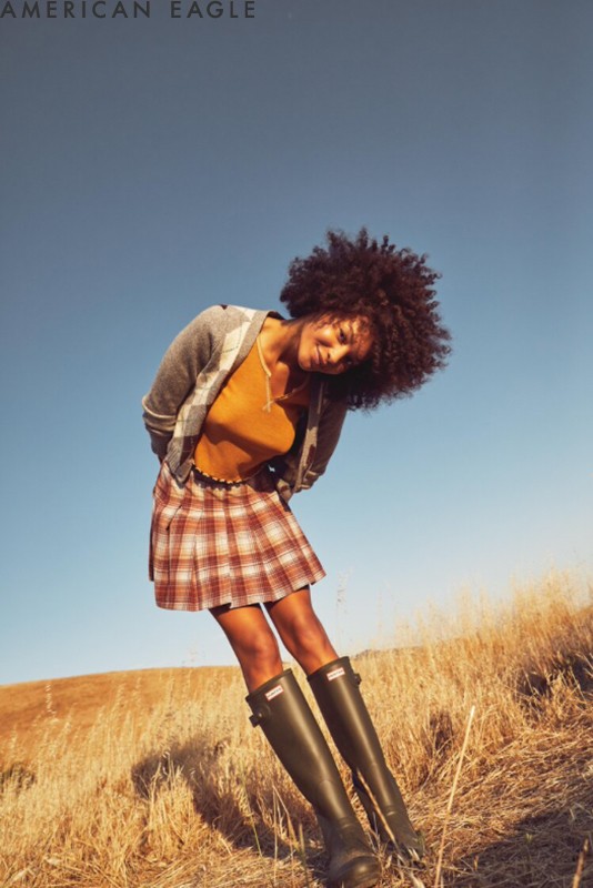 A young woman with curly hair in an AE plaid skirt, sweater and Hunter boots poses in a golden, grassy field under a blue sky.