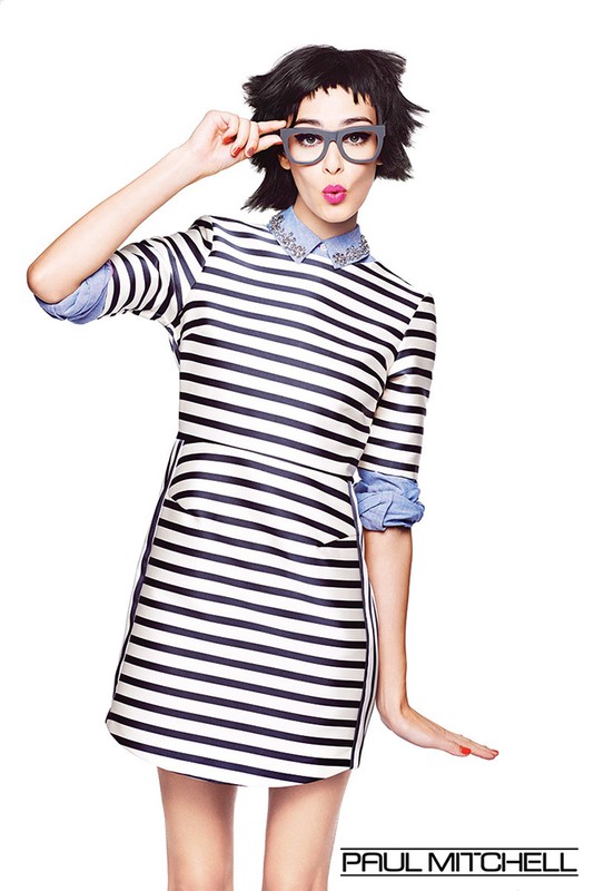 A fashionable adult female model with a dark wig, and striped dress poses in a photo studio wearing glasses. Beauty and fashion editorial.