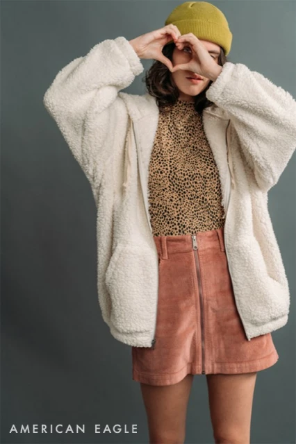 A woman poses in a sherpa jacket and corduroy skirt, heart-shaped hands framing her face, set against a gray studio backdrop.