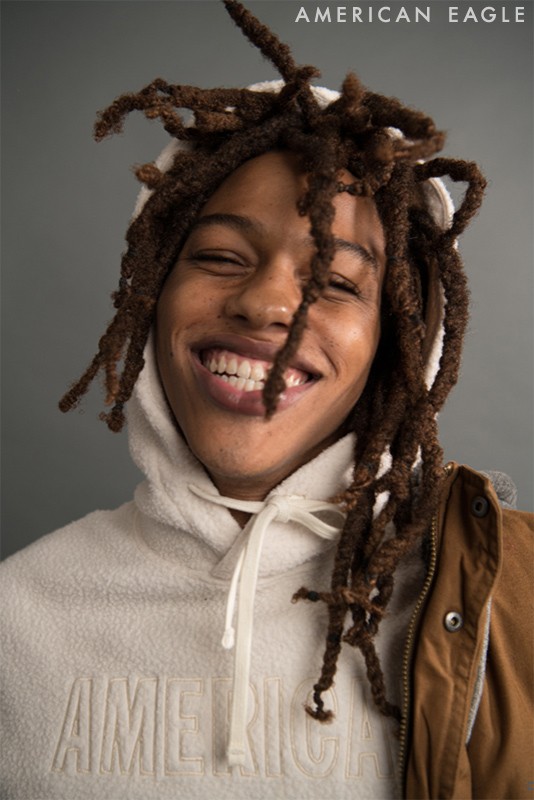 Close-up portrait of a smiling young Black man with dreadlocks, wearing a white hoodie and brown jacket, against a grey backdrop.