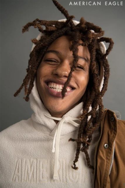 Close-up portrait of a smiling young Black man with dreadlocks, wearing a white hoodie and brown jacket, against a grey backdrop.