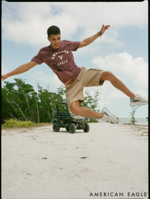 Young man jumping mid-air in front of a small black ATV on a sandy path near the beach on a sunny day, wearing a t-shirt and shorts.