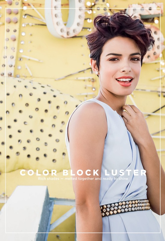 Portrait of a woman with short brown hair wearing a light blue dress, standing in front of a yellow patterned background.