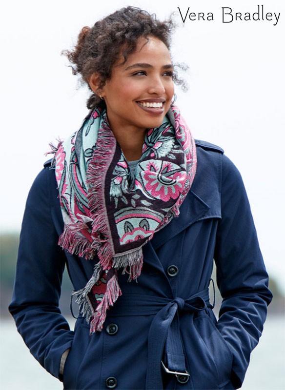 A smiling woman models a fringed floral scarf and navy trench coat for Vera Bradley against a blurred outdoor background.
