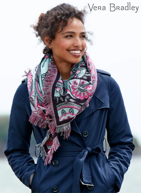 A smiling woman models a fringed floral scarf and navy trench coat for Vera Bradley against a blurred outdoor background.
