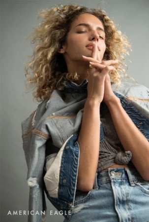 Curly-haired woman with eyes closed holding hands together in denim jacket and jeans against a grey backdrop