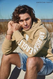 Young man with wavy brown hair wearing a tan hoodie and denim shorts, seated on a sandy beach.