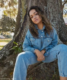 Woman wearing denim jacket and jeans seated in front of a large tree trunk in Los Angeles