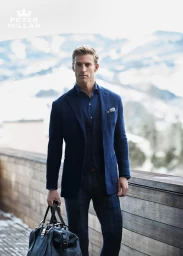 Man wearing a blue blazer and vest holding a matching duffel bag outdoors against a snowy mountain backdrop.