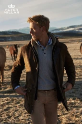Man wearing a brown suede jacket over a grey polo and khakis outdoors in a dry landscape with mountains and horses.