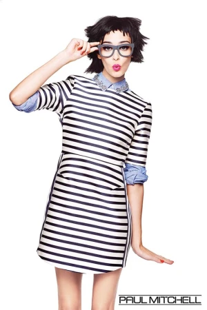 Woman in striped dress and thick grey glasses holding her frames while posing against a white background.