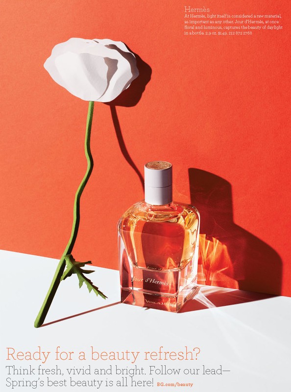 Perfume bottle and white paper flower on a white surface against a reddish-orange backdrop, showcasing minimalist beauty and floral fragrance.
