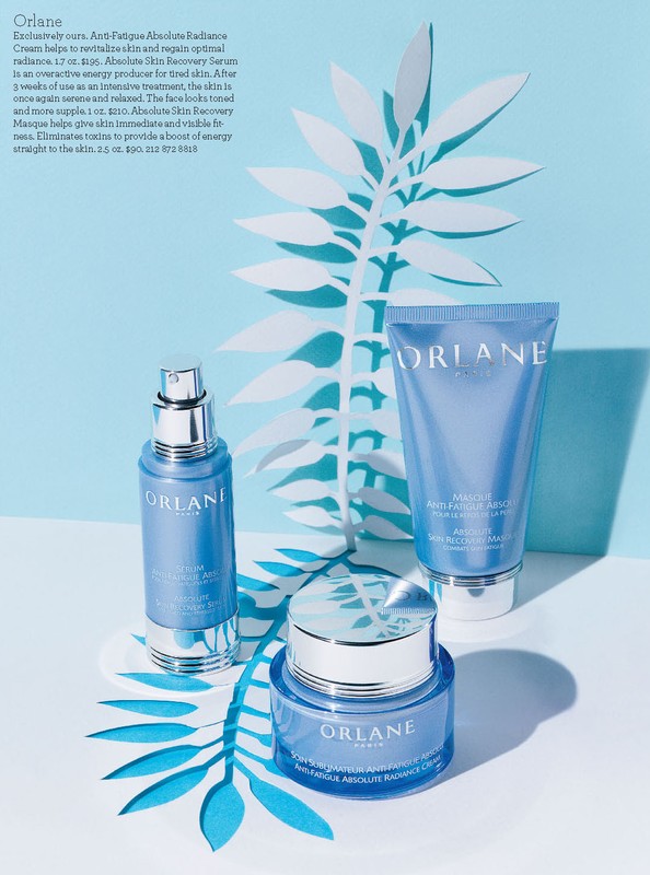 Three Orlane skincare products against a light blue backdrop with paper leaf cutouts, showcasing serum, cream, and lotion for beauty.
