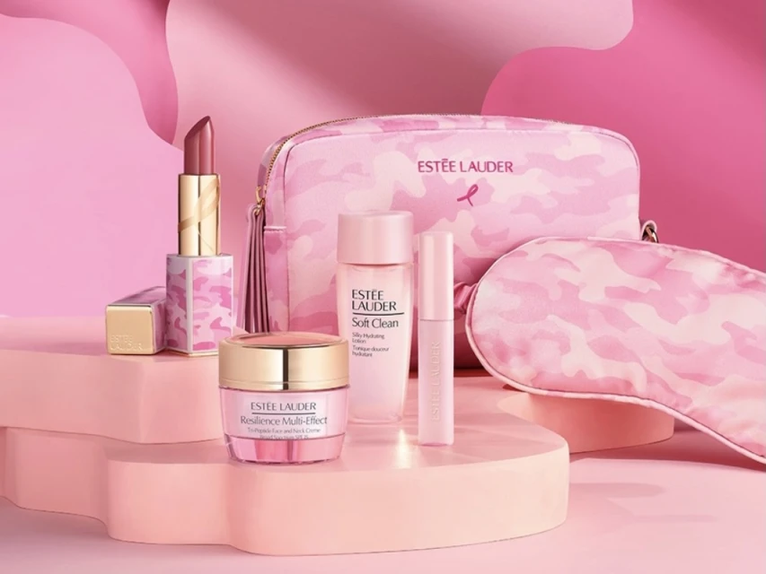 Estee Lauder makeup and skincare products including lipstick, night mask, moisturizer, and cleanser on a pink multi-tiered platform.
