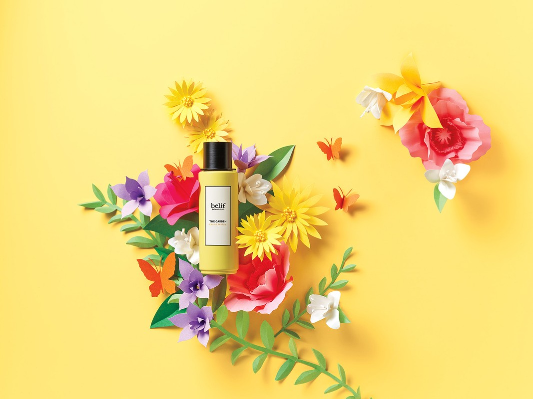 Still life of skincare product with yellow bottle on yellow background surrounded by colorful paper flowers and paper butterflies.