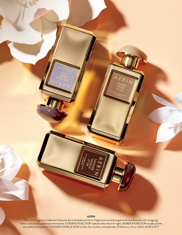 Three gold AERIN perfume bottles are arranged on a peach surface surrounded by white paper flowers in a beauty product advertisement.
