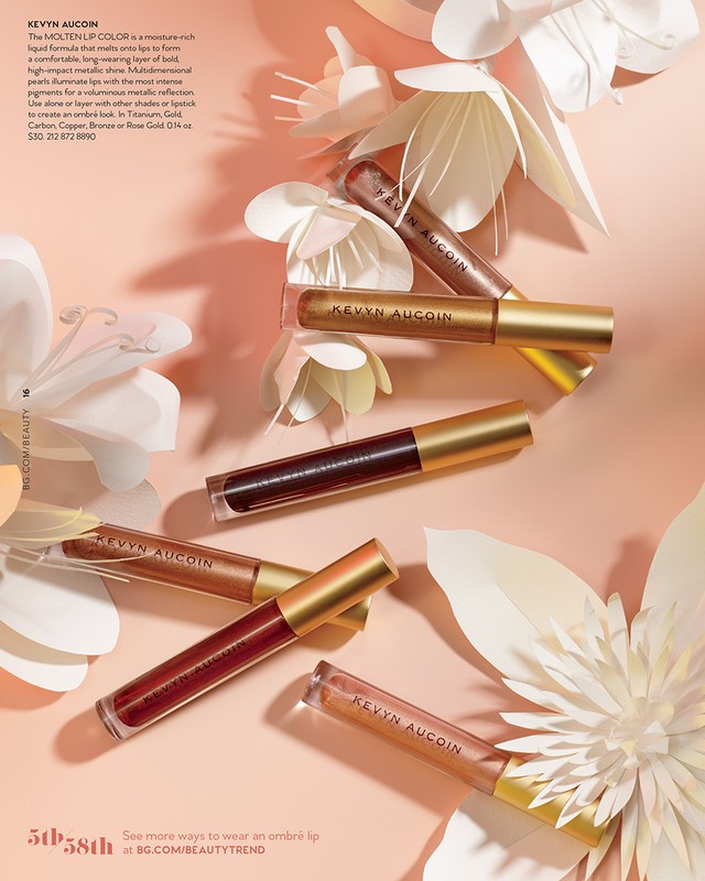 Beauty editorial showcasing an arrangement of Kevyn Aucoin lip gloss tubes in varying shades, interspersed with delicate paper flower decorations.
