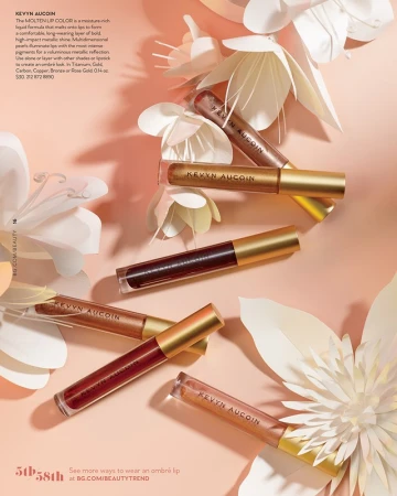 Beauty editorial showcasing an arrangement of Kevyn Aucoin lip gloss tubes in varying shades, interspersed with delicate paper flower decorations.