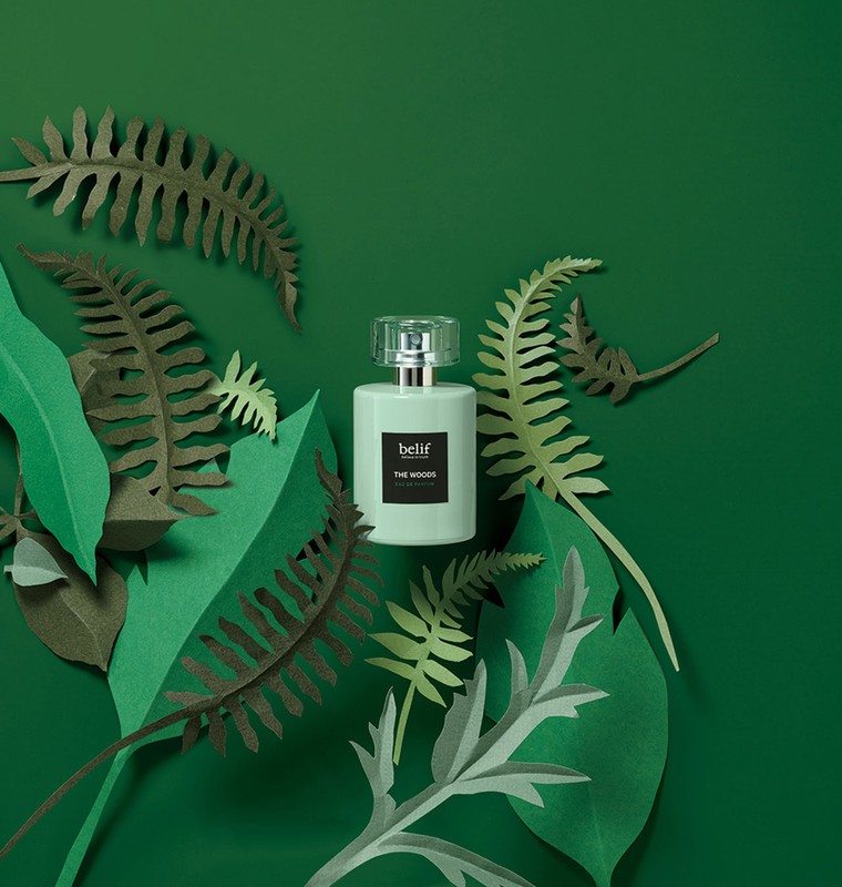 Belif perfume bottle nestled among green paper fern fronds on a green background in a beauty still-life studio product shot.