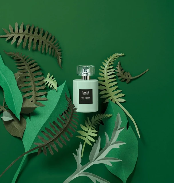 Belif perfume bottle nestled among green paper fern fronds on a green background in a beauty still-life studio product shot.