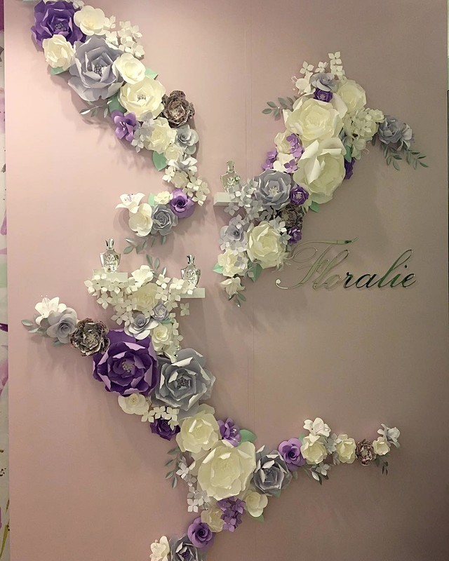 Elegant floral wall decoration featuring white, grey, and purple paper flowers arranged in a sweeping design with a 'Floralie' script.