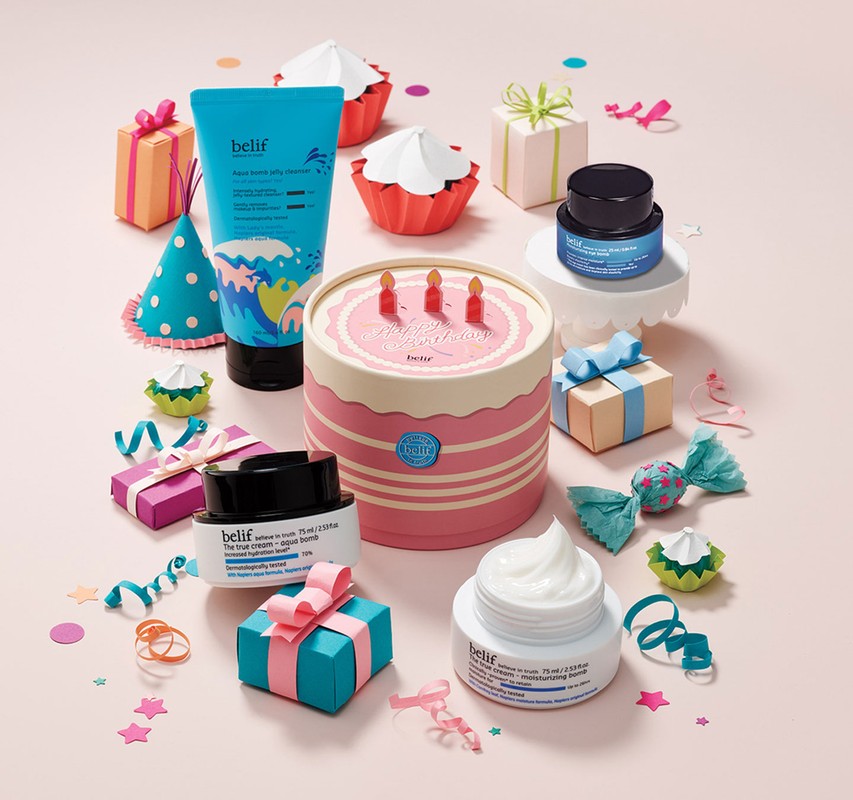 Cosmetic products from Belif are arranged with decorative gift boxes, paper cupcakes, and confetti on a pink background for a product shot.