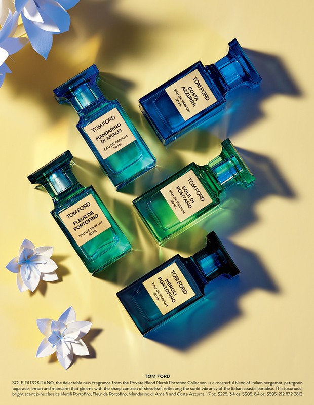 Five colorful glass Tom Ford perfume bottles are arranged on a soft yellow backdrop with white paper flower accents, product photography.