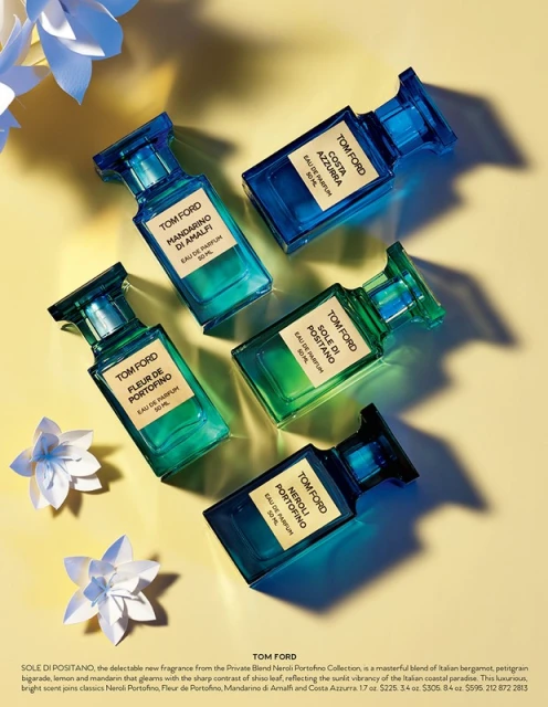 Five colorful glass Tom Ford perfume bottles are arranged on a soft yellow backdrop with white paper flower accents, product photography.