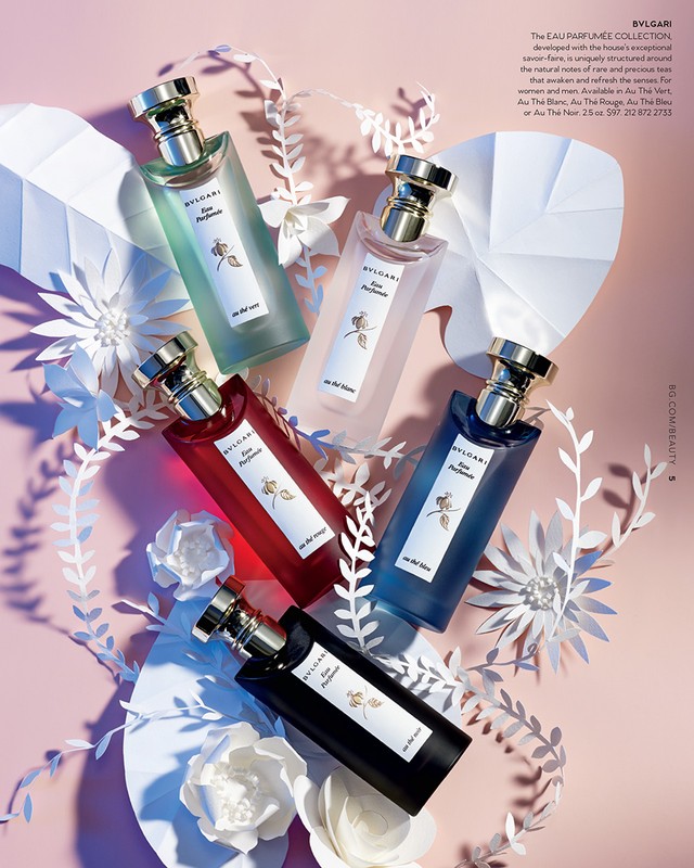 Five BVLGARI perfume bottles in varied colors, surrounded by paper foliage and flowers, artfully arranged on a soft pink background.
