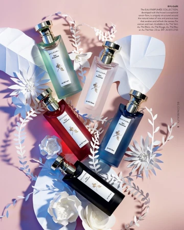 Five BVLGARI perfume bottles in varied colors, surrounded by paper foliage and flowers, artfully arranged on a soft pink background.